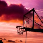 "Basketball hoop silhouette against a dramatic sunset sky with purple and orange clouds"