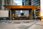 Modern Apple Store with glass façade and glowing Apple logo surrounded by tall city buildings at sunset.