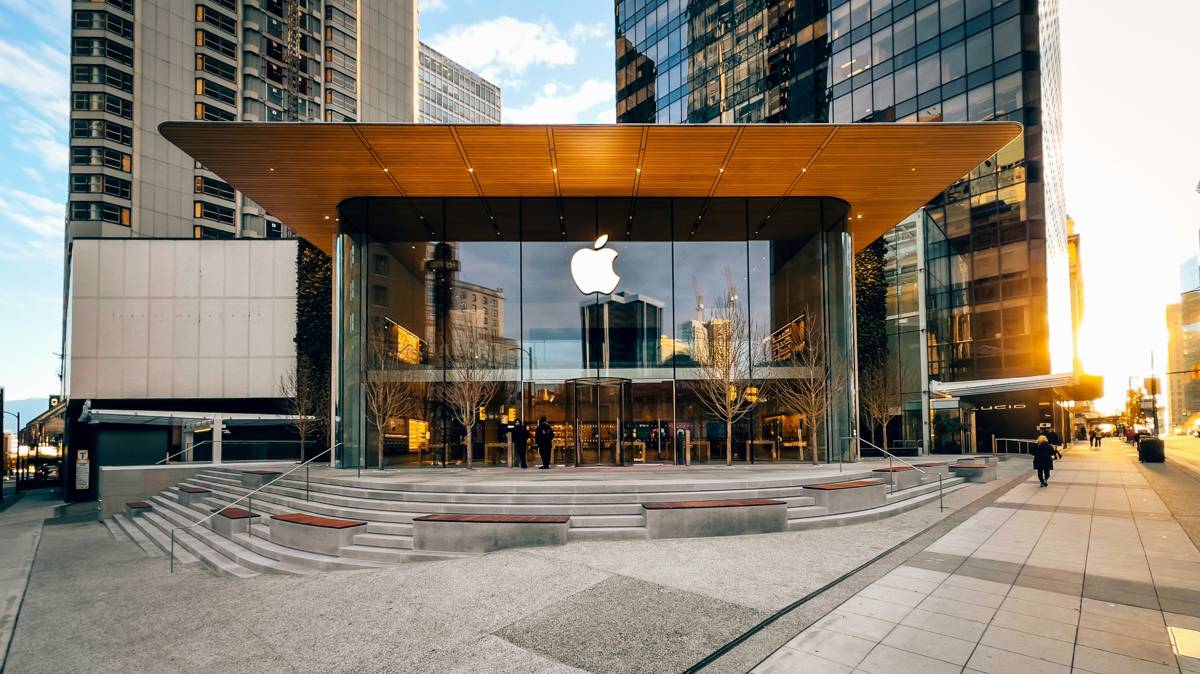 Modern Apple Store with glass façade and glowing Apple logo surrounded by tall city buildings at sunset.
