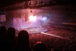 A massive concert crowd cheering under vibrant stage lights and fireworks during a live music performance in a packed arena.