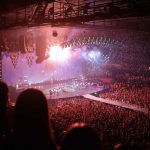 A massive concert crowd cheering under vibrant stage lights and fireworks during a live music performance in a packed arena.