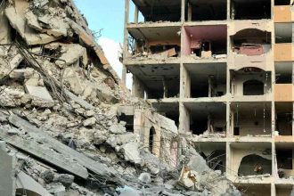 Destroyed residential building in Gaza after airstrikes, with rubble and collapsed concrete filling the street below.