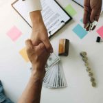 Two people shaking hands over a business deal with money, keys, and contract documents on the table.