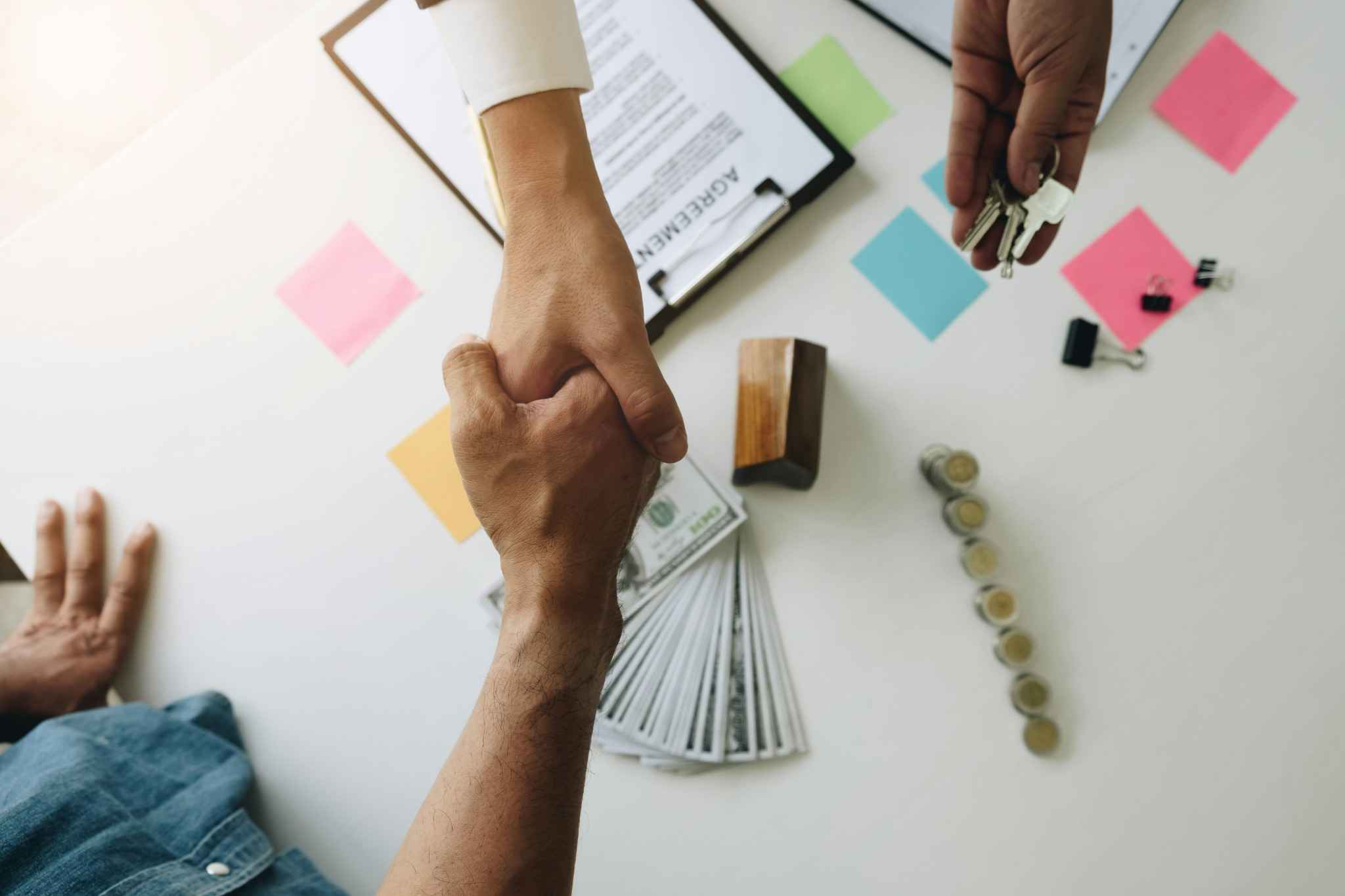 Two people shaking hands over a business deal with money, keys, and contract documents on the table.
