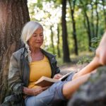 Senior woman relaxing outdoors, sitting under a tree and reading a book in the forest.