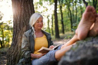 Senior woman relaxing outdoors, sitting under a tree and reading a book in the forest.