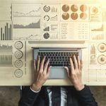 Business professional working on a laptop with financial graphs and data analytics visualizations.