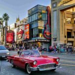 A classic red convertible driving along Hollywood Boulevard, passing by the Hard Rock Cafe and Dolby Theatre under clear blue skies.