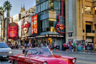 A classic red convertible driving along Hollywood Boulevard, passing by the Hard Rock Cafe and Dolby Theatre under clear blue skies.