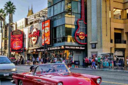 A classic red convertible driving along Hollywood Boulevard, passing by the Hard Rock Cafe and Dolby Theatre under clear blue skies.