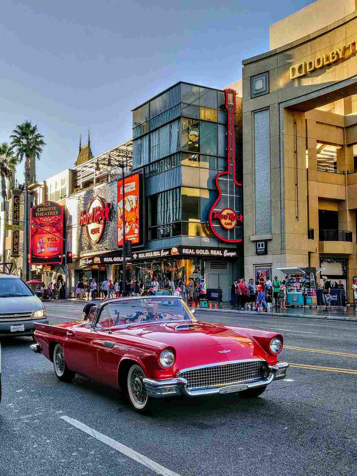 A classic red convertible driving along Hollywood Boulevard, passing by the Hard Rock Cafe and Dolby Theatre under clear blue skies.