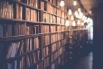 Rows of books on shelves in a library with hanging light bulbs.