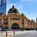Flinders Street Station in Melbourne, Australia, with trams, cars, and people in front of the historic building under a clear blue sky.