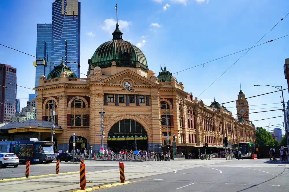 Flinders Street Station in Melbourne, Australia, with trams, cars, and people in front of the historic building under a clear blue sky.