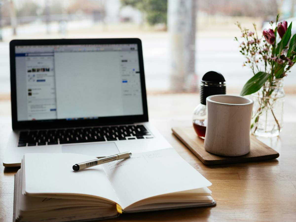 Laptop, notebook, pen, and coffee cup on a wooden desk by the window.