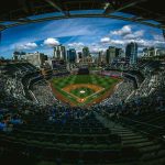 Panoramic view of a packed baseball stadium under a bright blue sky with a city skyline in the background.
