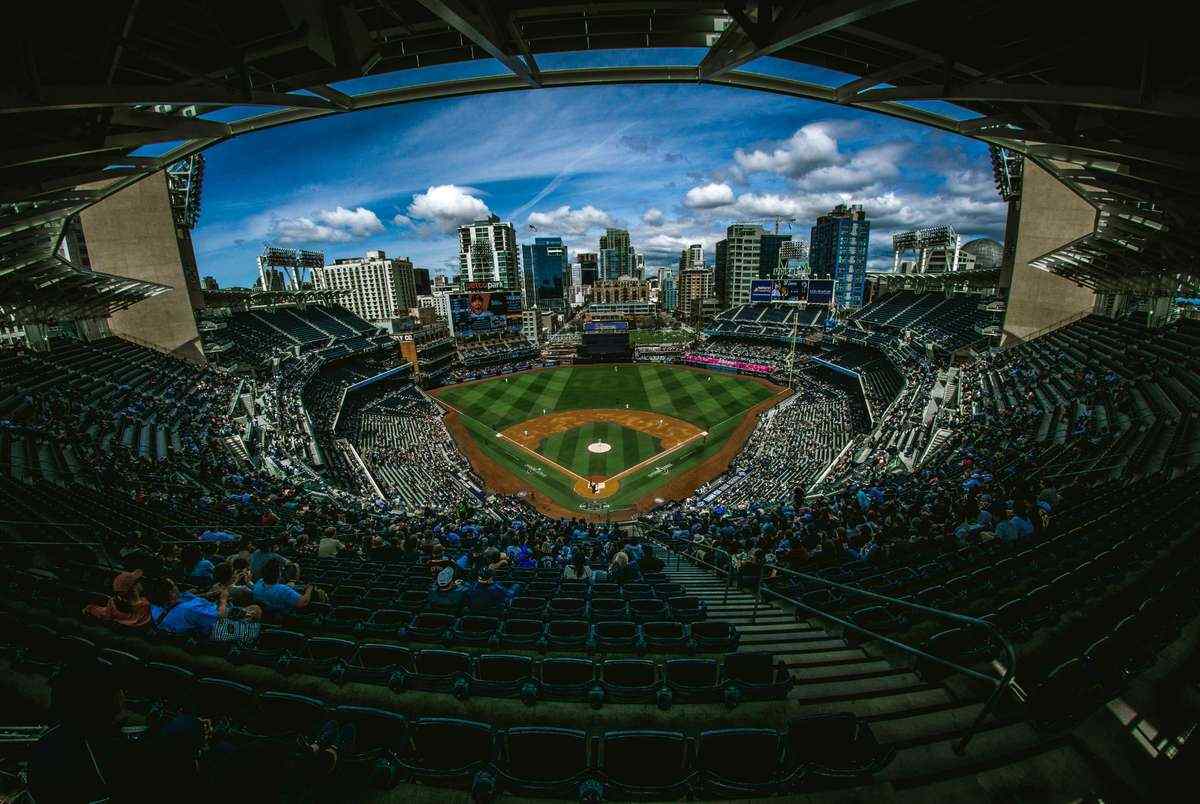 Panoramic view of a packed baseball stadium under a bright blue sky with a city skyline in the background.