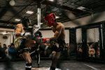 Two fighters in protective gear exchanging kicks inside an MMA cage during a live match.