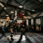 Two fighters in protective gear exchanging kicks inside an MMA cage during a live match.