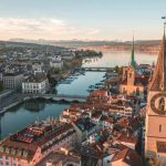 A stunning aerial view of Zurich, Switzerland, showing church spires, historic rooftops, and Lake Zurich framed by distant snow-capped mountains.