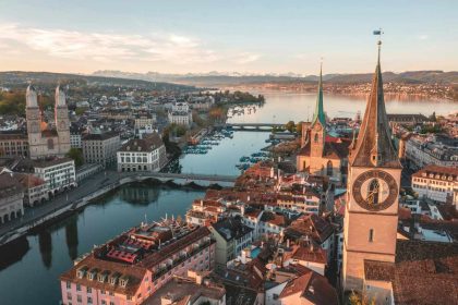 A stunning aerial view of Zurich, Switzerland, showing church spires, historic rooftops, and Lake Zurich framed by distant snow-capped mountains.