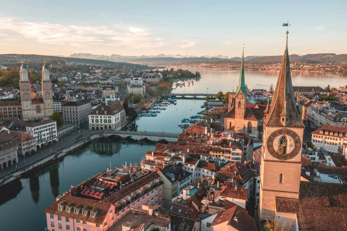 A stunning aerial view of Zurich, Switzerland, showing church spires, historic rooftops, and Lake Zurich framed by distant snow-capped mountains.