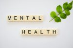 The word “Mental Health” spelled with Scrabble tiles placed neatly beside a green leaf on a white background.