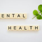 The word “Mental Health” spelled with Scrabble tiles placed neatly beside a green leaf on a white background.