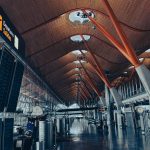 Modern airport departure hall with bright lighting, large display boards, and travelers walking through the spacious terminal.